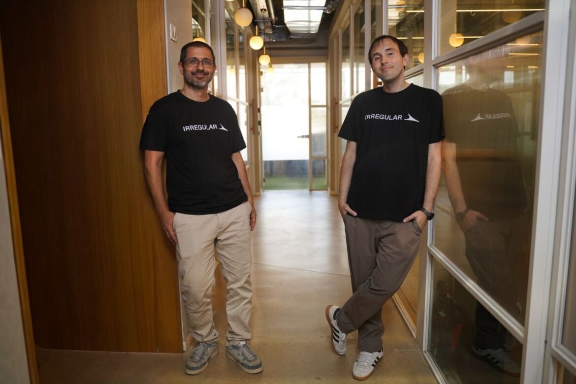 The two co-founders stand abreast in an office hallway wearing black logo t-shirts.