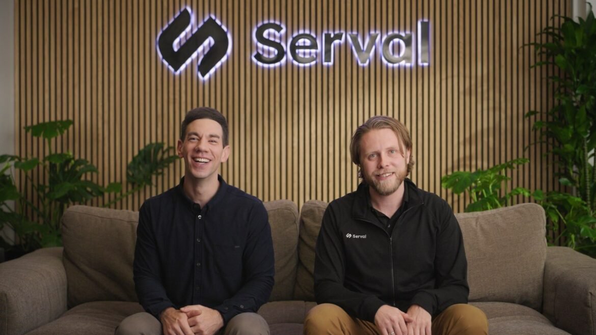 Two caucasian men sit on a couch with a company logo behind them.