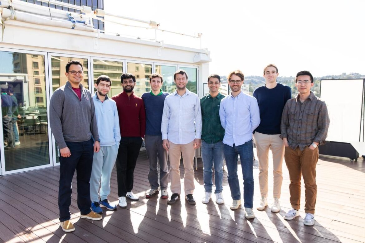 A group of nine men stand on a sunlit balcony.