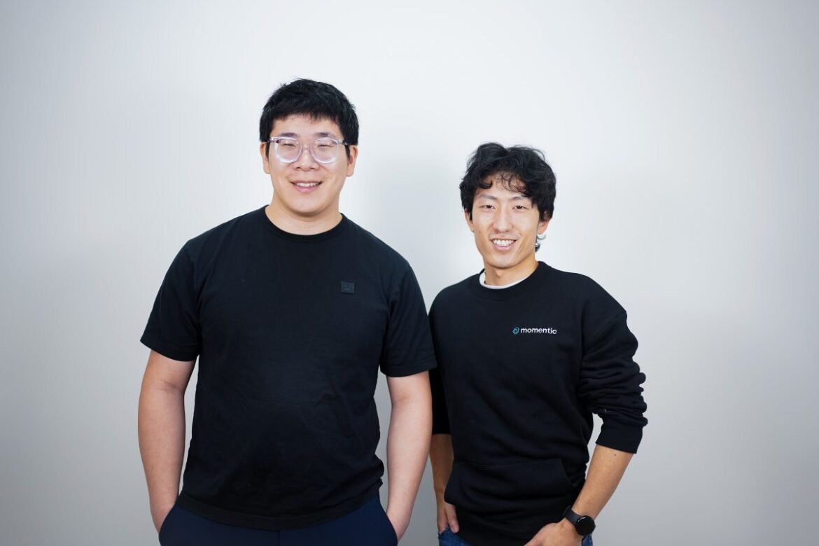 Two men in black t-shirts stand against a gray background.